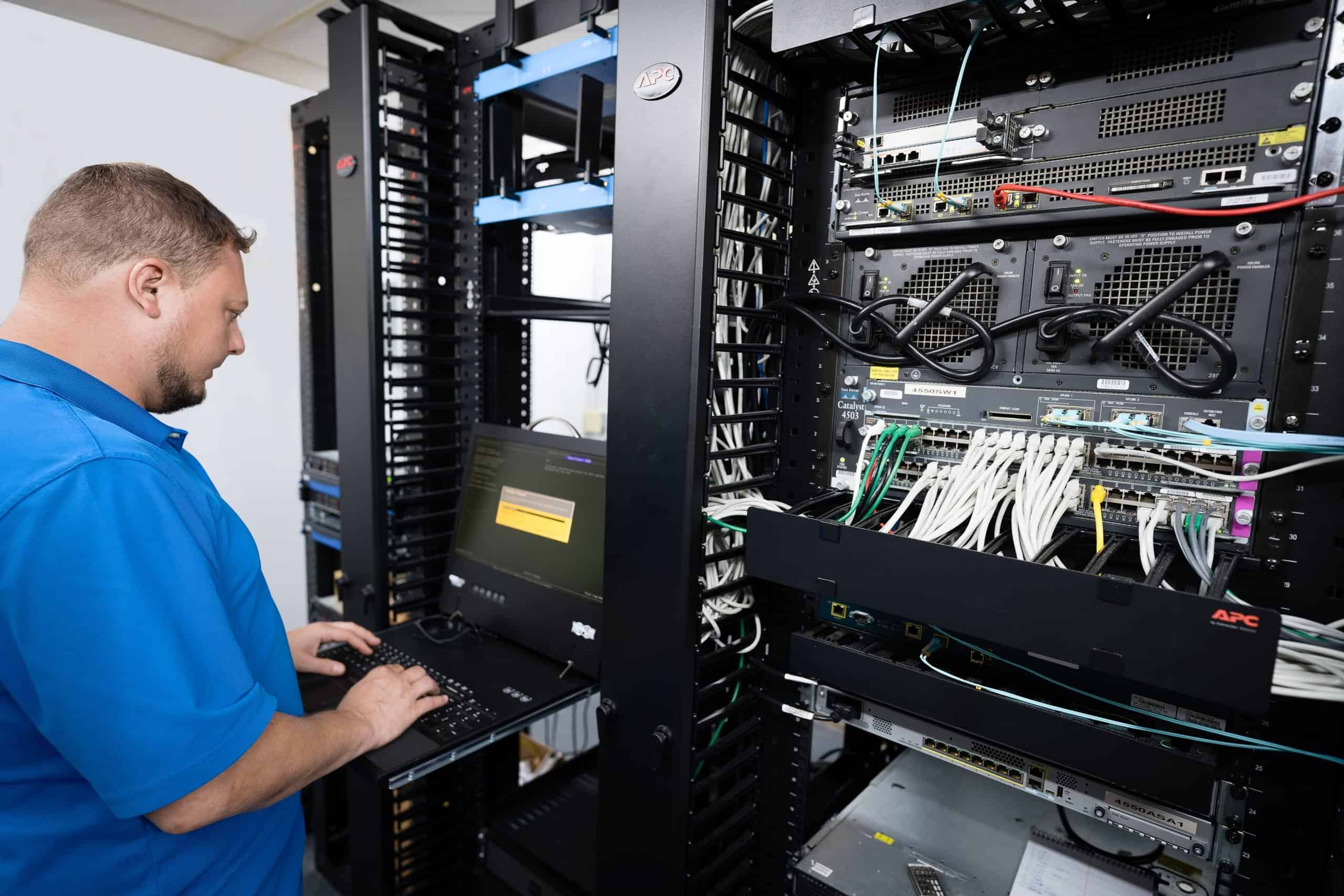 IT technician working at a keyboard beside a server rack with patch panels