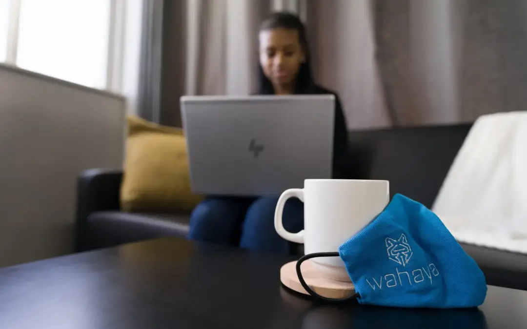 A blue face mask and white mug on a table with a woman working on a laptop in the background.