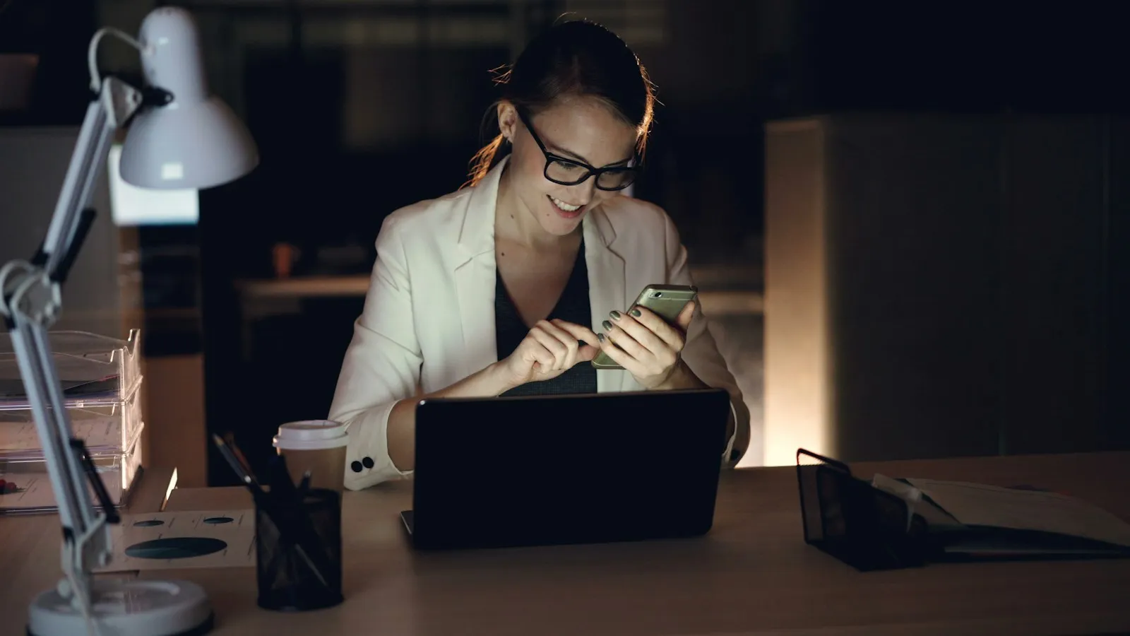 Woman checking her phone at a laptop in a dim office